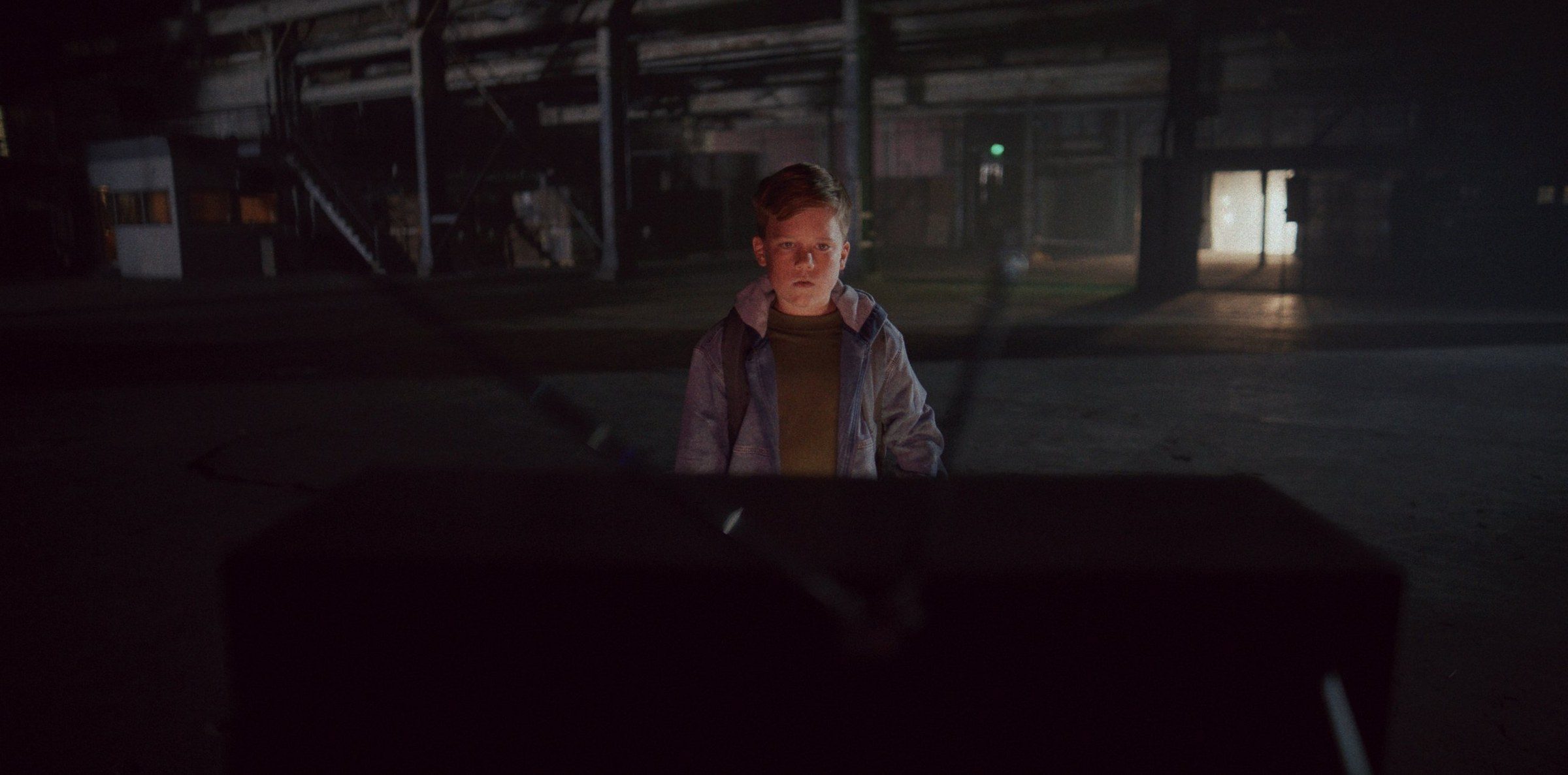 A boy lit by a TV in a vast dark warehouse
