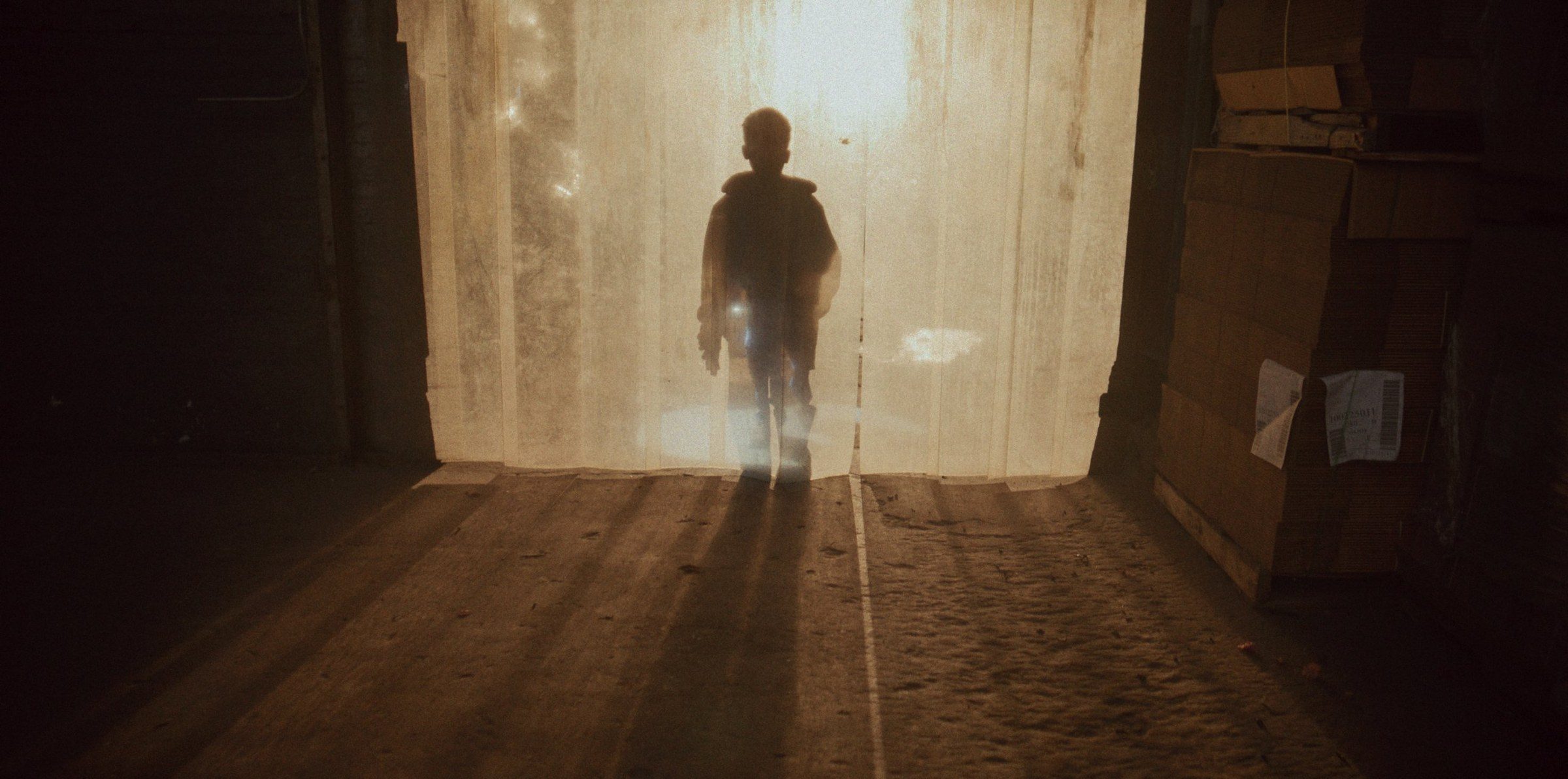 A boy silhouetted against a glowing plastic curtain in a warehouse doorway