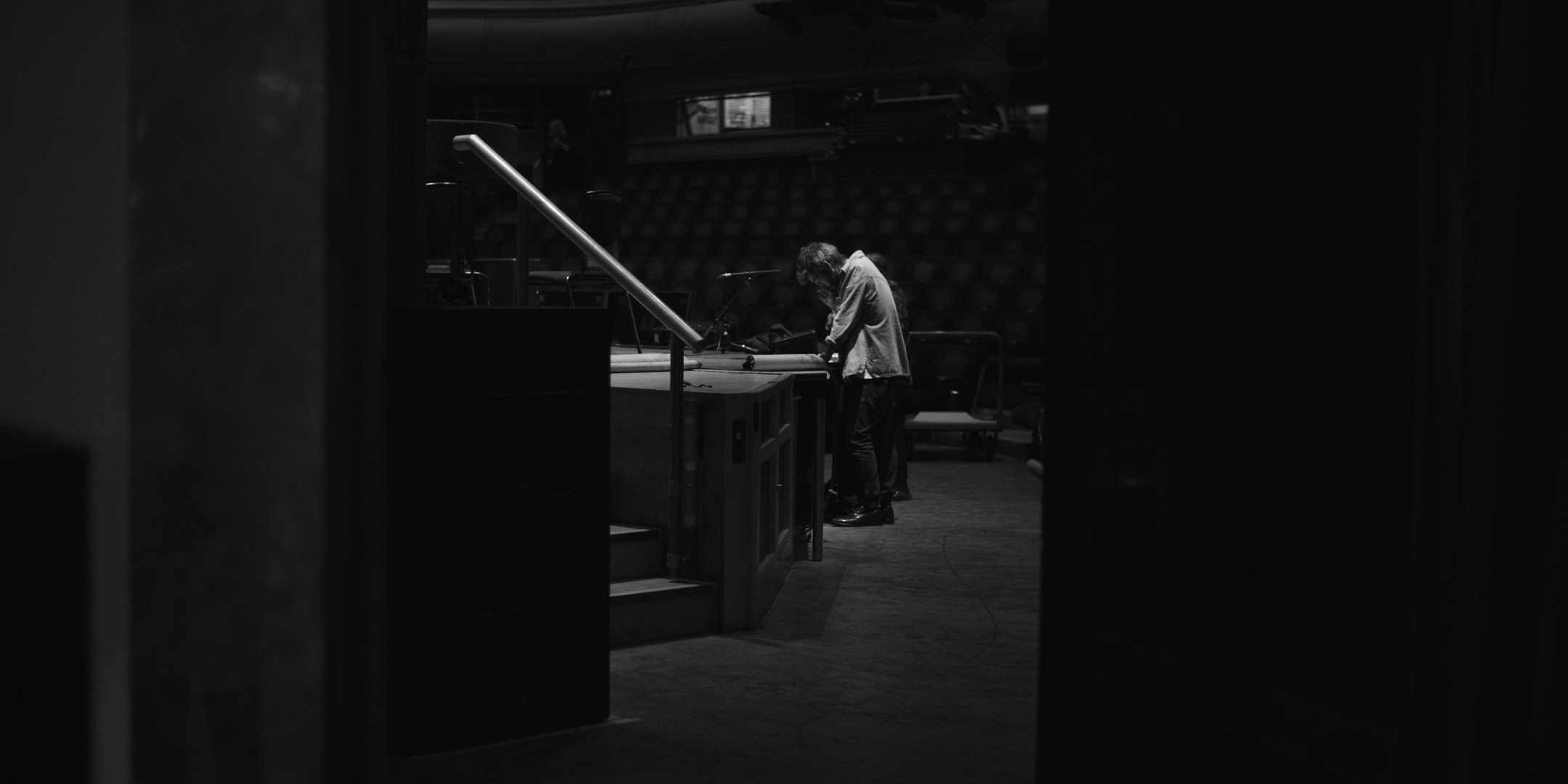 A distant figure at a piano seen through a dark backstage doorway