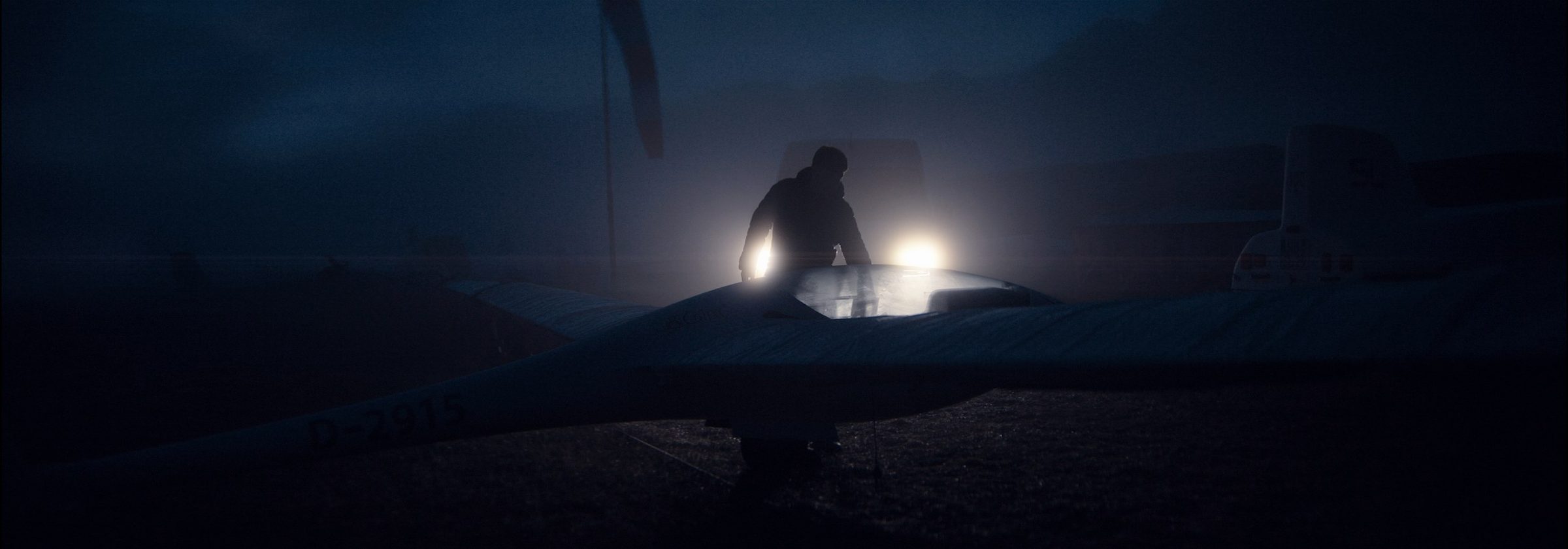 A figure at a glider on a dark airfield, two lamps glowing