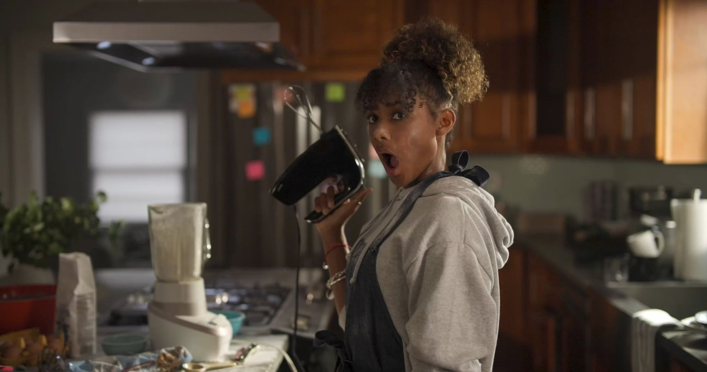 A girl in a kitchen holding up a hand mixer, mouth open in shock