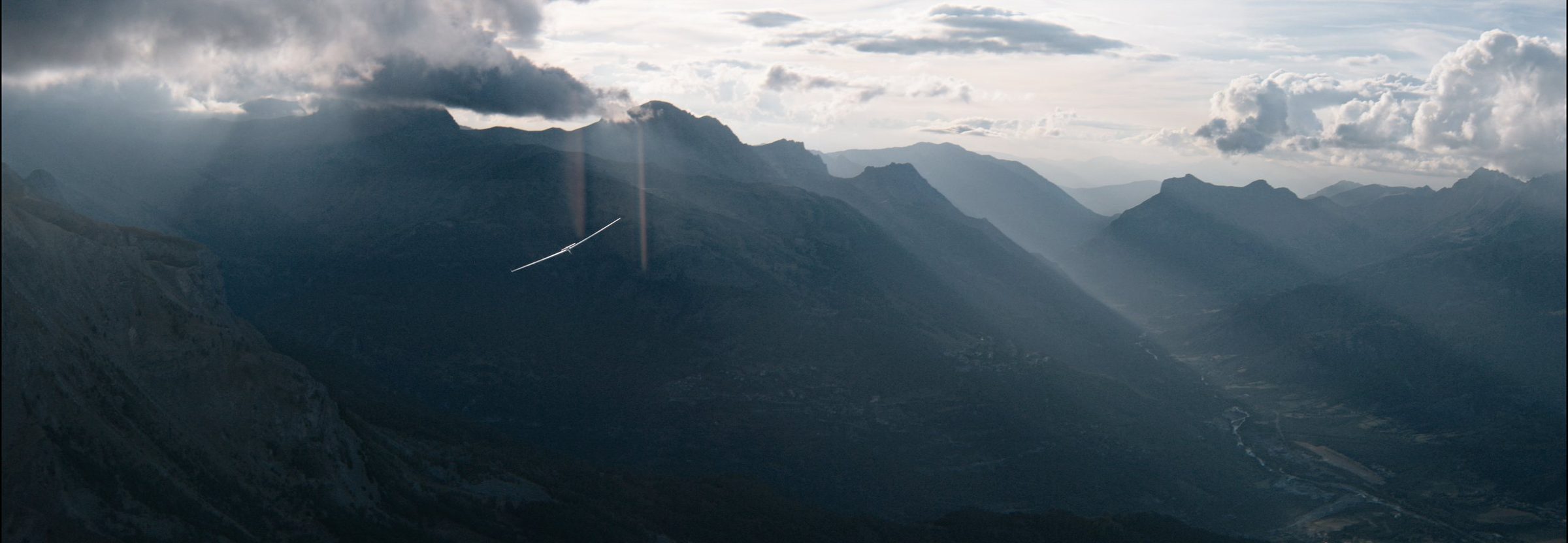 A glider tiny against a vast alpine valley, light raking through cloud