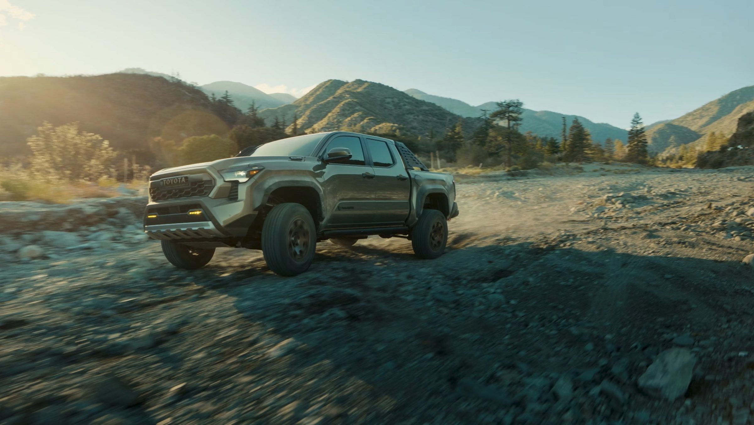 A green Toyota Tacoma kicking up dust on a mountain riverbed at sunset