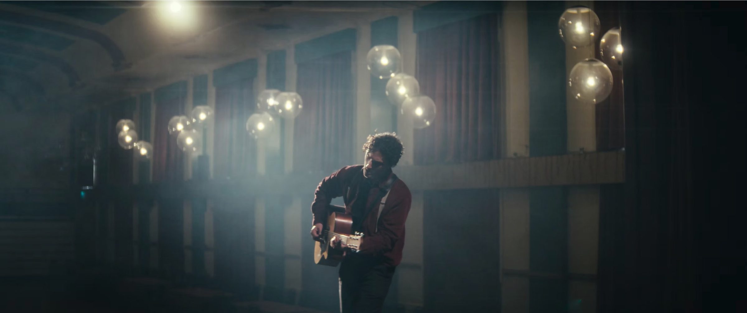 A guitarist playing in an empty ballroom under hanging globe lights