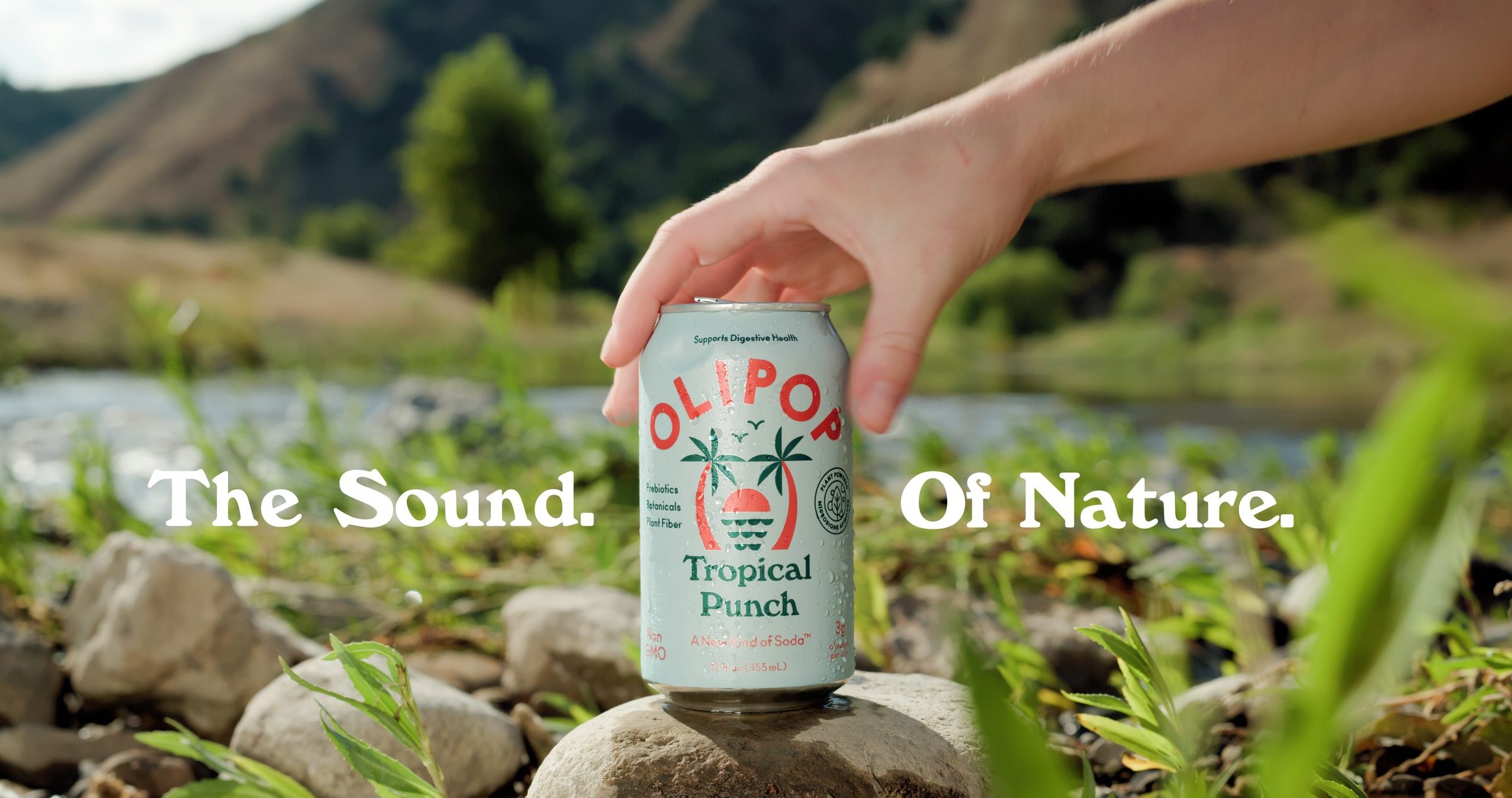 A hand setting a can of Olipop on a river stone, hills behind
