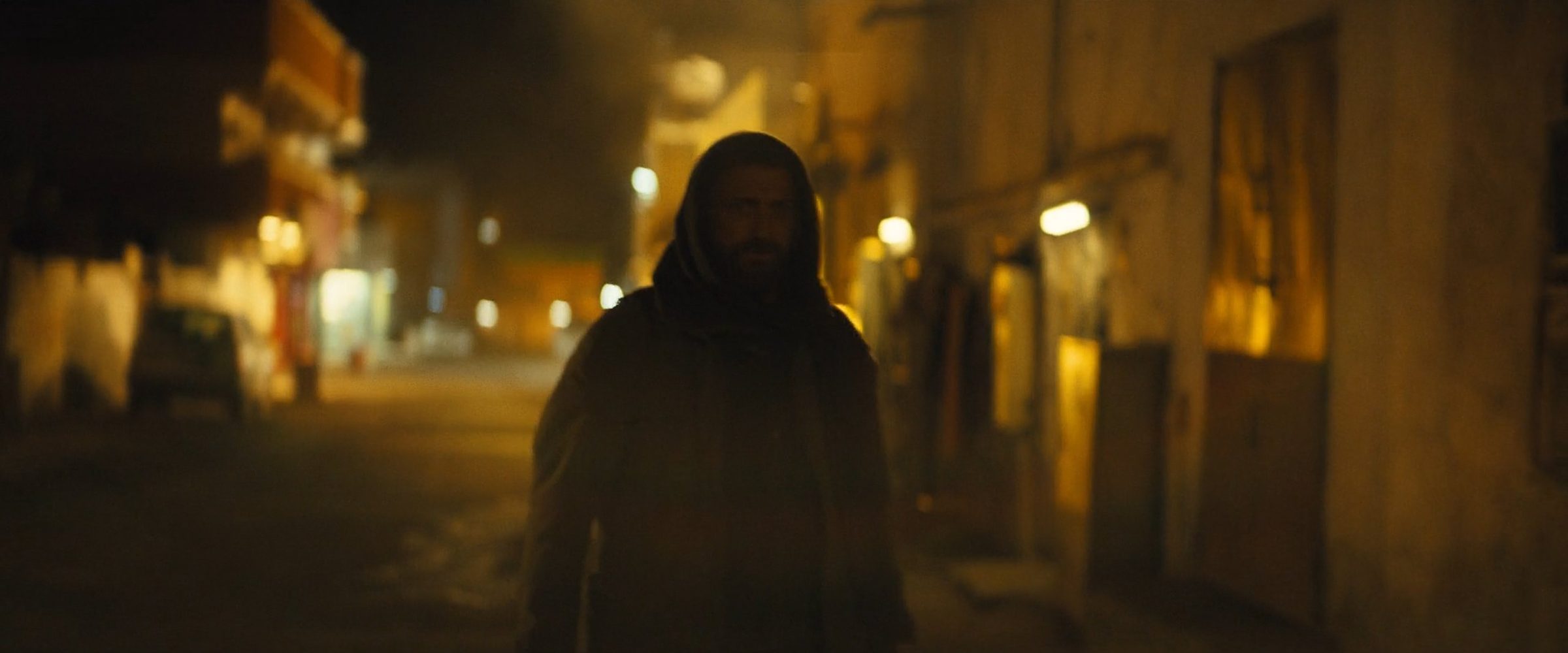 A hooded man walking down a lamplit night street