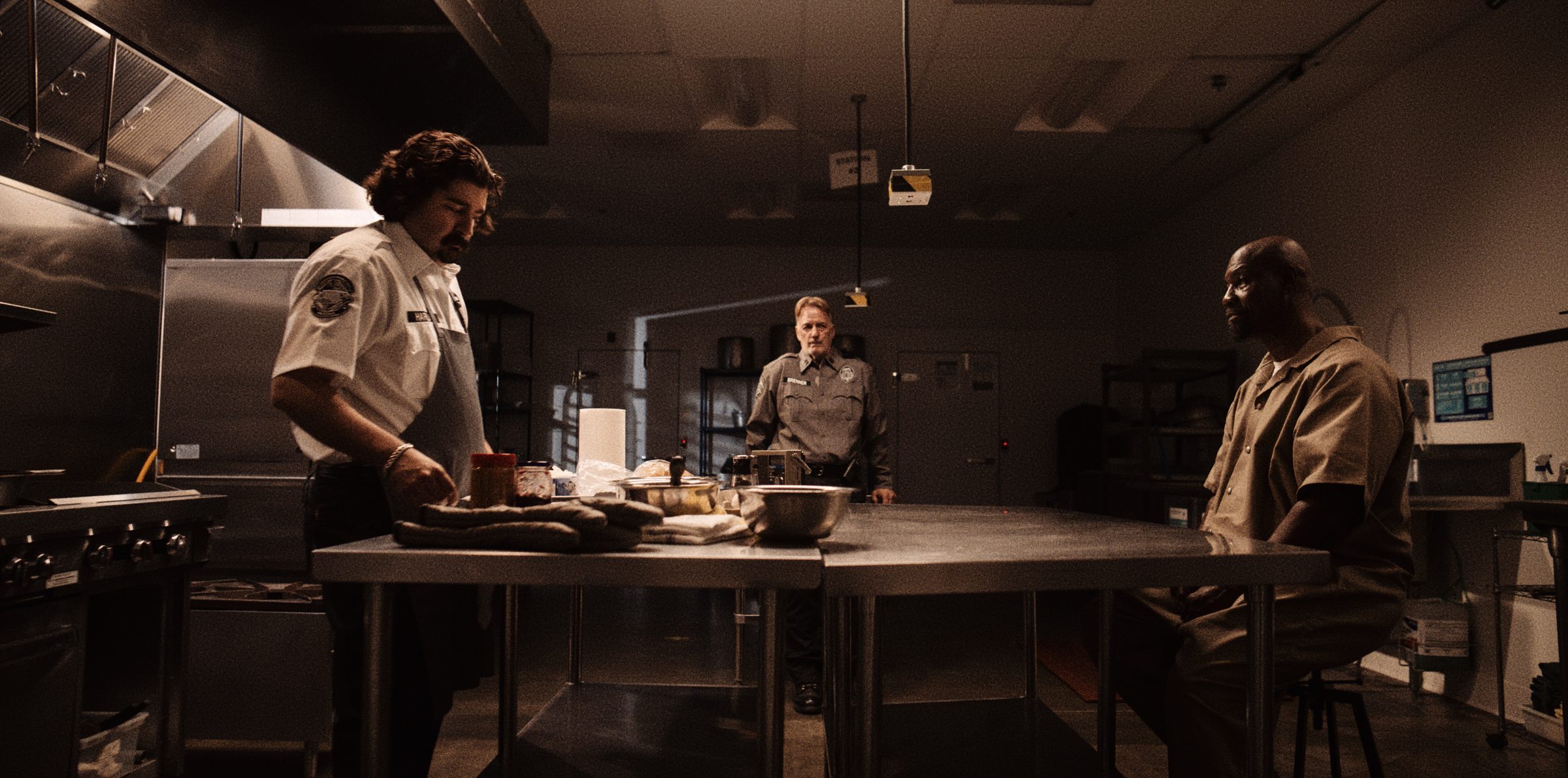 A prison kitchen at night: a cook, an officer and an inmate in pools of low light