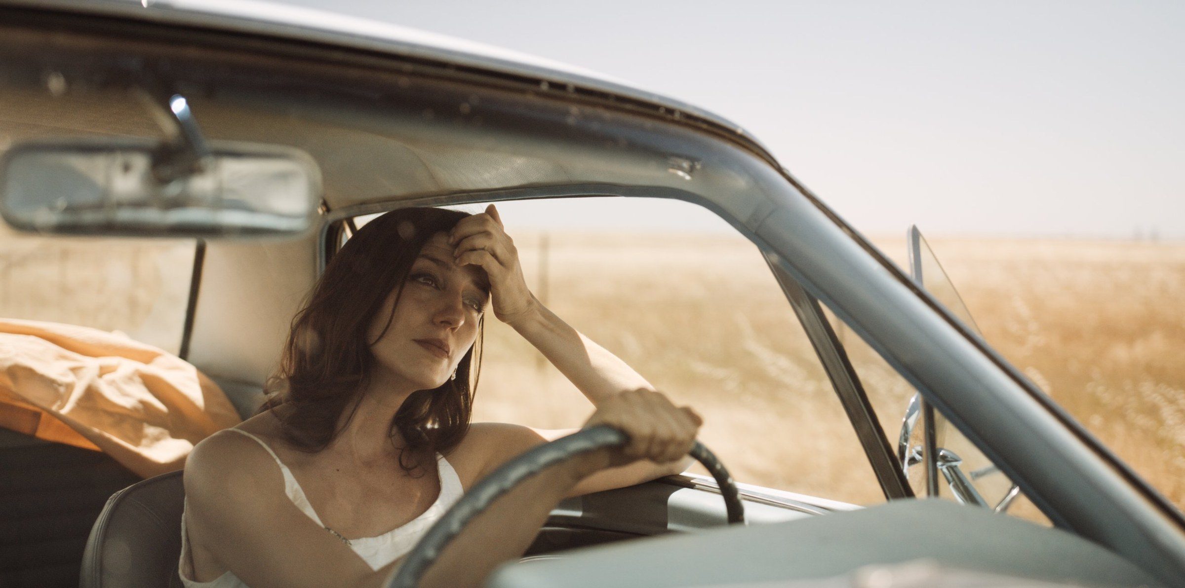A woman at the wheel, hand to her forehead, wheat fields behind