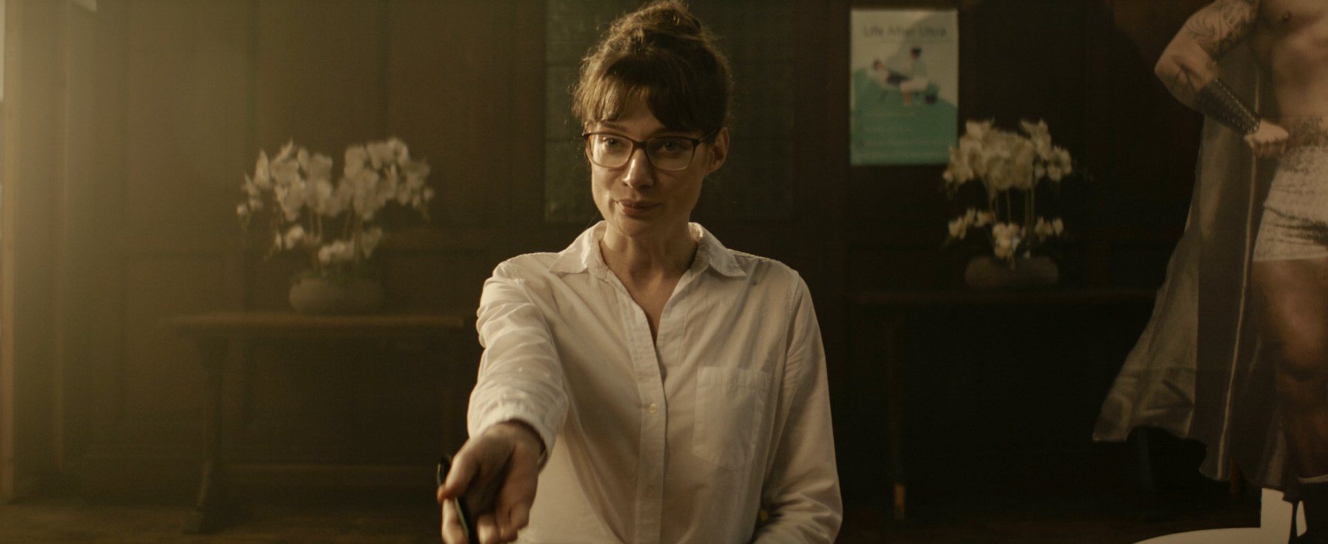 A woman in a white shirt pointing a pen across a dim wood-panelled office