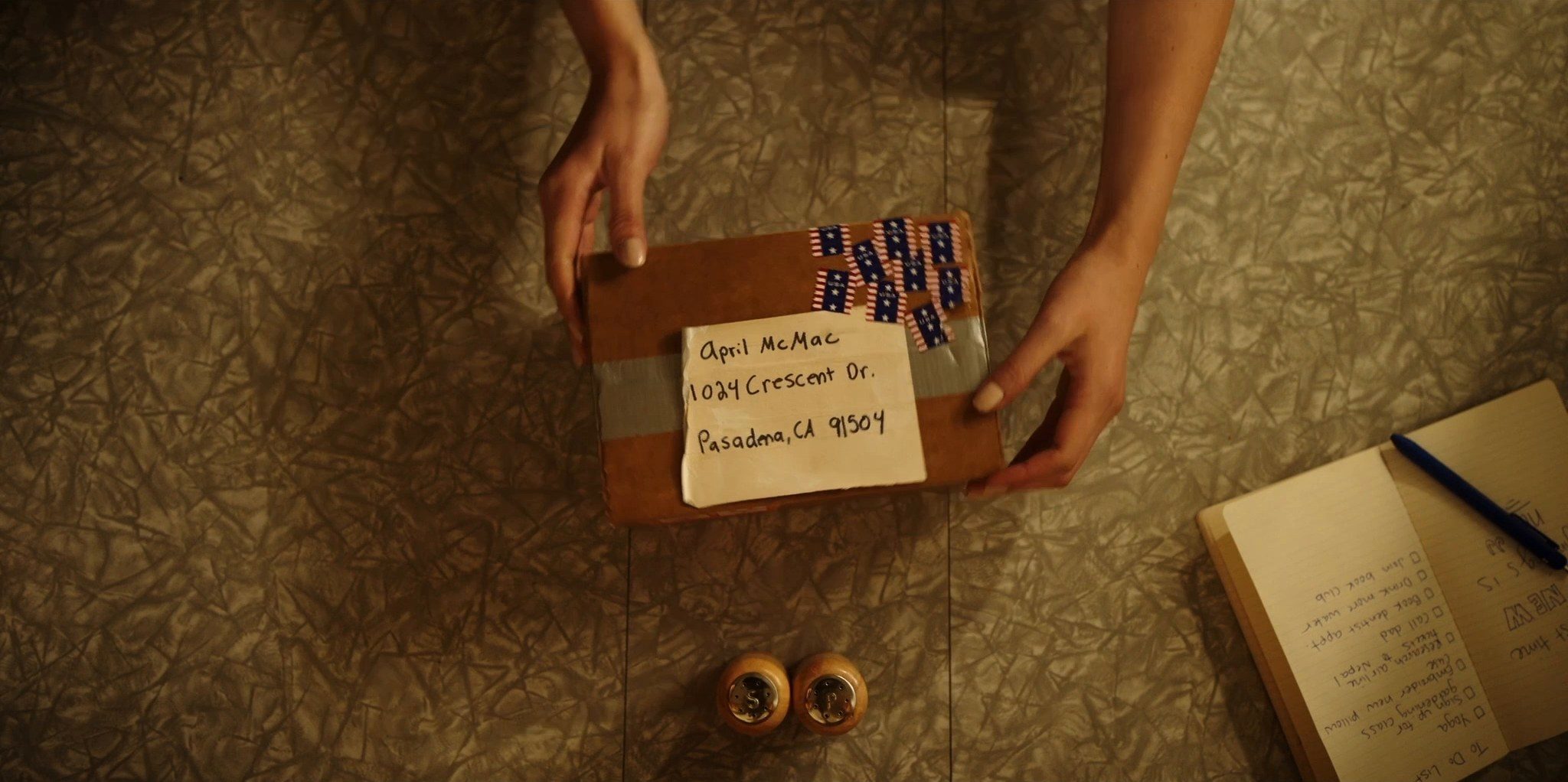 Hands placing a taped parcel on a patterned floor beside a notebook